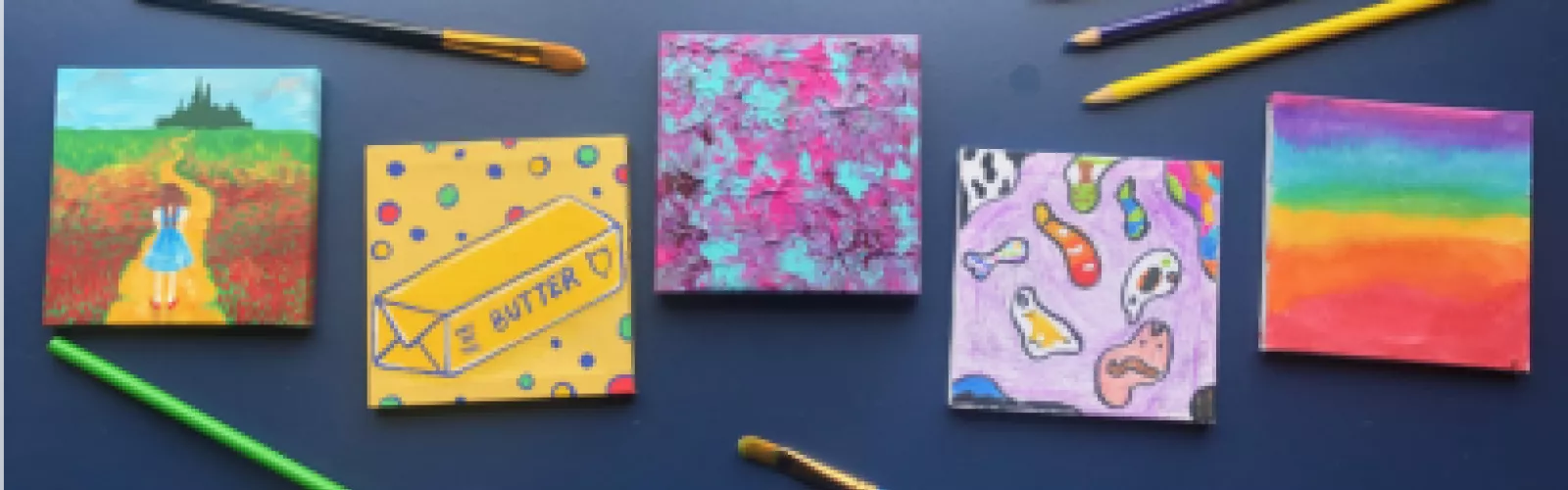 Small square painted canvasses lined up in a row surrounded by scattered paint brushes and colored pencils.